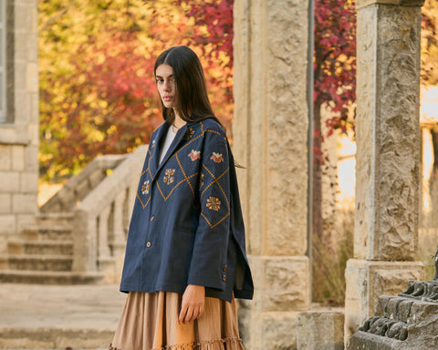 Woman in a blue embroidered jacket and beige skirt standing in an outdoor setting with stone architecture and autumn foliage.